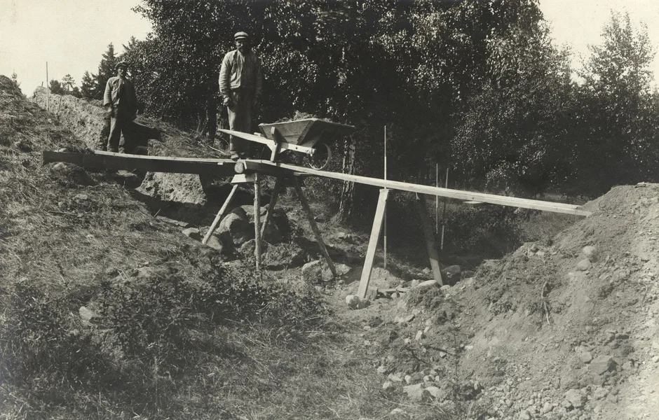 Två arkeologer ståendes på en bro av plankor ovanför en arkeologisk utgrävningsplats